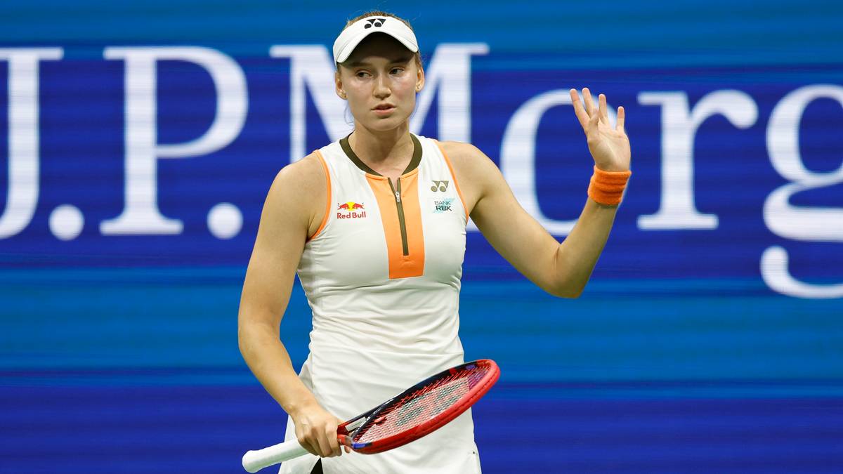 Tenisistka w białej koszulce sportowej z pomarańczowymi wstawkami, białej spódniczce i czapce z daszkiem, z pomarańczową opaską na prawym nadgarstku, trzymająca rakietę tenisową w lewej dłoni. Wznosi prawą rękę, machając palcami, gestykulując do publiczności. Na jej koszulce widoczne logo Red Bulla i numer 22. W tle widać niebieskie i białe pasy oraz napisy 'J.P.Morgan'.