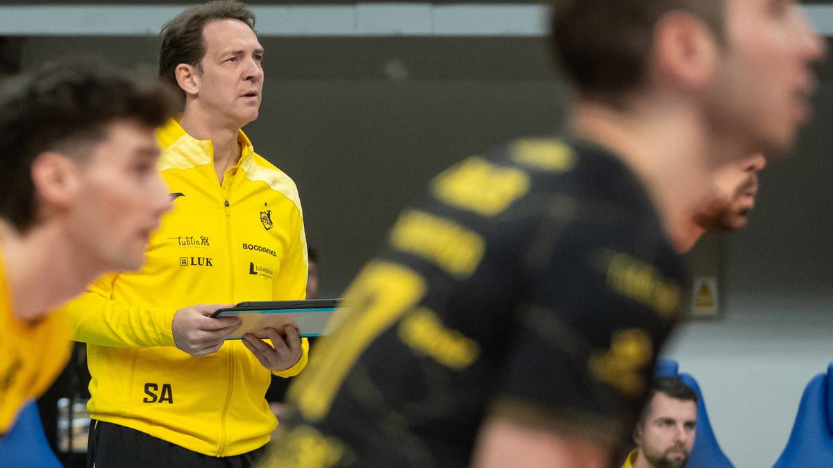 Trener siatkówki w żółtej kurtce z tabletem w ręku obserwuje grę.