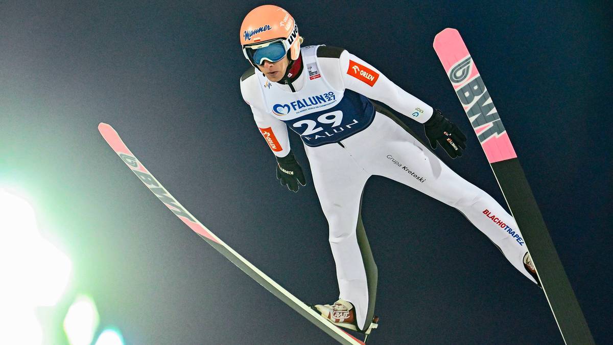 Skoczek narciarski w locie, w białym kombinezonie i pomarańczowym kasku, na tle ciemnego nieba