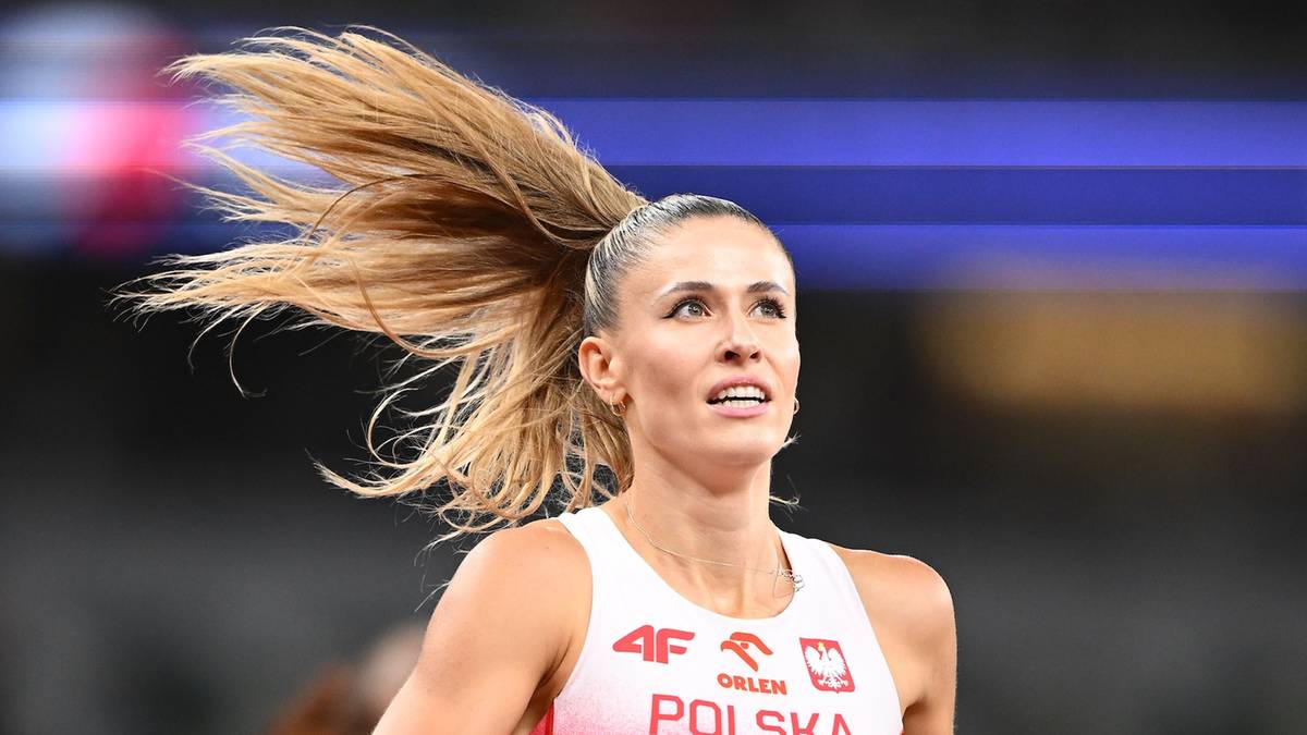 Portret lekkoatletki Natalii Bukowieckiej podczas zawodów. Ma związane w wysoki kucyk blond włosy, które rozwiewają się na wietrze. Ubrana jest w białą koszulkę sportową z czerwonymi elementami i napisem "POLSKA".