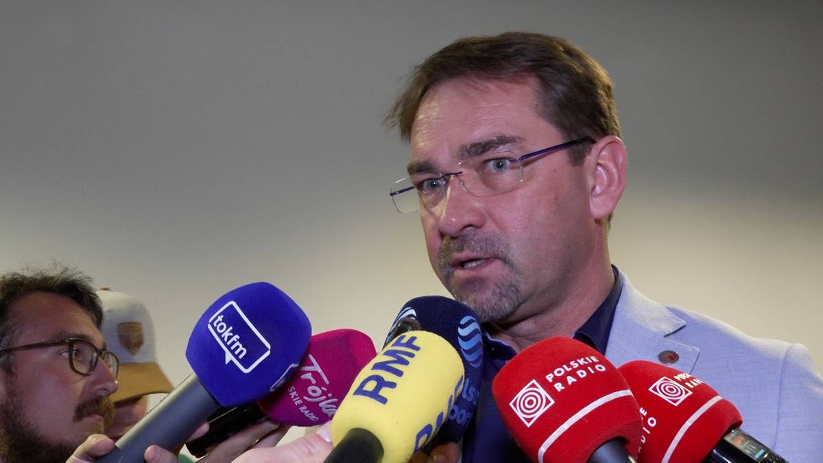 Mężczyzna w okularach i jasnej marynarce przemawia do grupy mikrofonów.