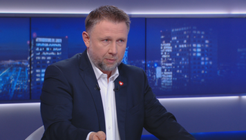 Mężczyzna w granatowym garniturze z białą koszulą i rozpiętym kołnierzykiem, siedzi przy stole i gestykuluje prawą ręką.