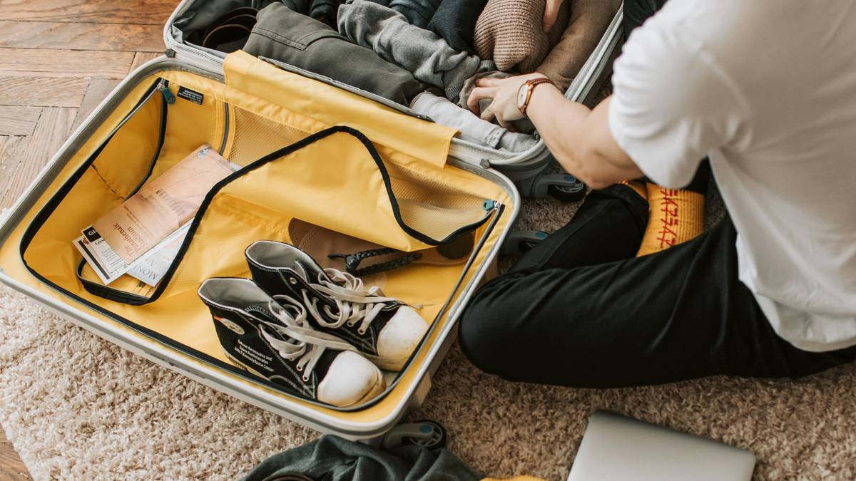 Osoba pakuje walizkę, obok leżą ubrania, buty i laptop.