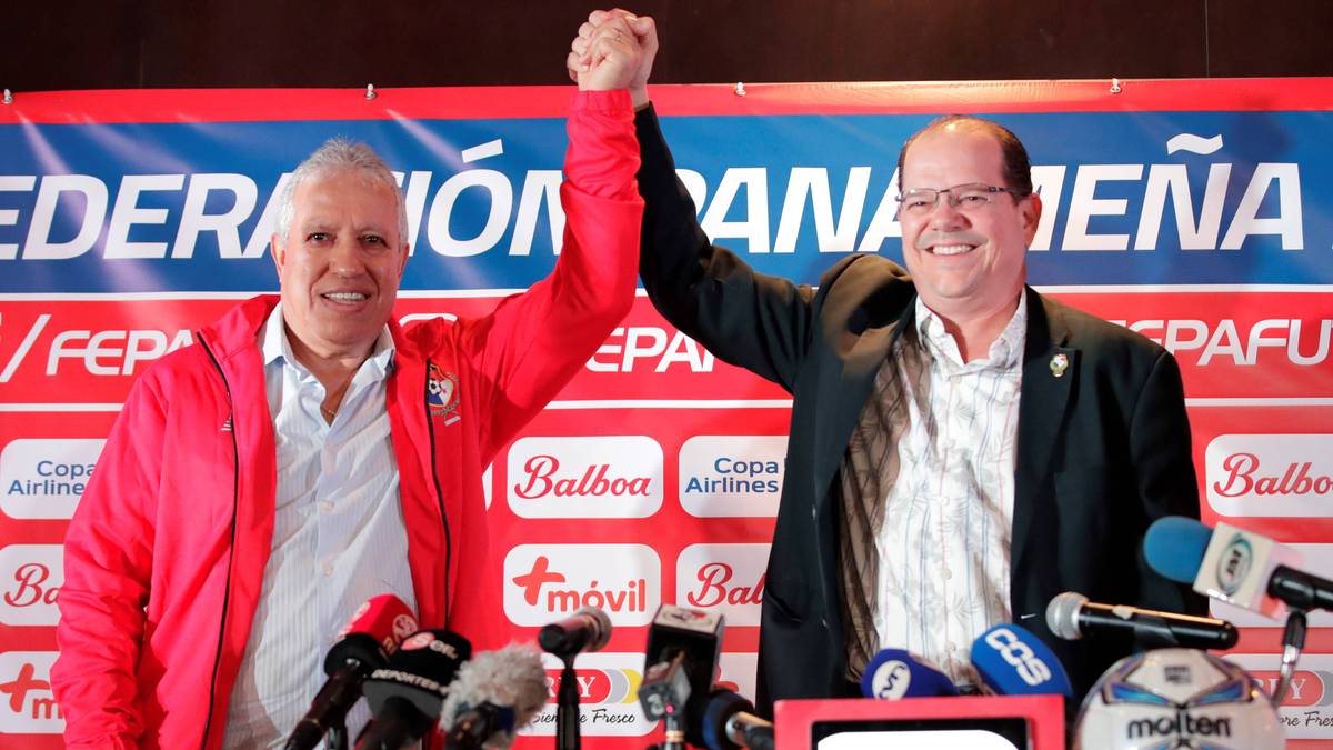 Dwóch mężczyzn, jeden w czerwonej kurtce, drugi w marynarce, pozuje do zdjęć za mównicą z mikrofonami, podczas gdy jeden z nich unosi pięść w geście zwycięstwa.