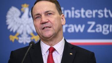 Portret mężczyzny w garniturze i krawacie, m&oacute;wiącego do mikrofonu.