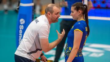 Trener w białej koszulce rozmawia z młodą siatkarką w niebieskim stroju sportowym, wskazując coś na tablecie.
