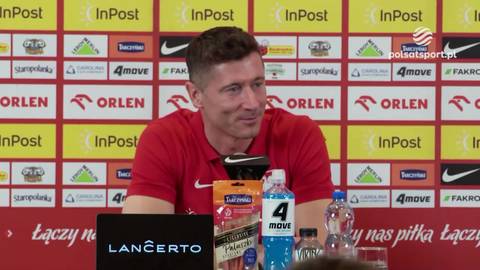 Robert Lewandowski w czerwonej koszulce reprezentacji Polski siedzi za stołem podczas konferencji prasowej. Za nim widoczne logotypy sponsor&oacute;w.