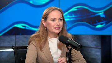 Kobieta w jasnym żakiecie siedzi przy mikrofonie, patrząc w stronę kamery.