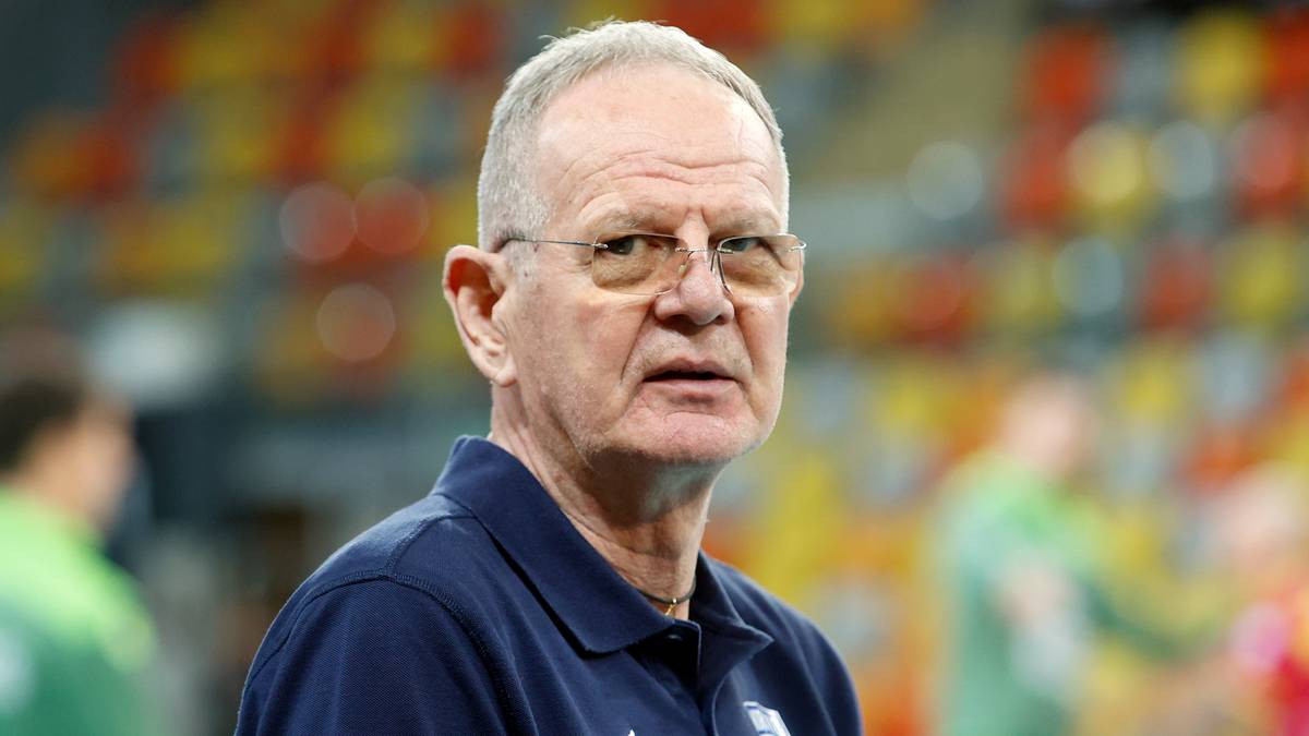 Portret starszego mężczyzny w okularach, ubranego w granatową koszulkę polo z niewielkim logo. Tło jest rozmyte i przedstawia trybuny z kolorowymi siedzeniami.