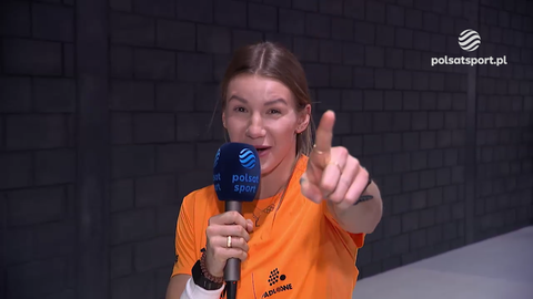 Adrianna Sułek-Schubert w pomarańczowej koszulce z mikrofonem Polsat Sport wskazuje palcem w stronę kamery.