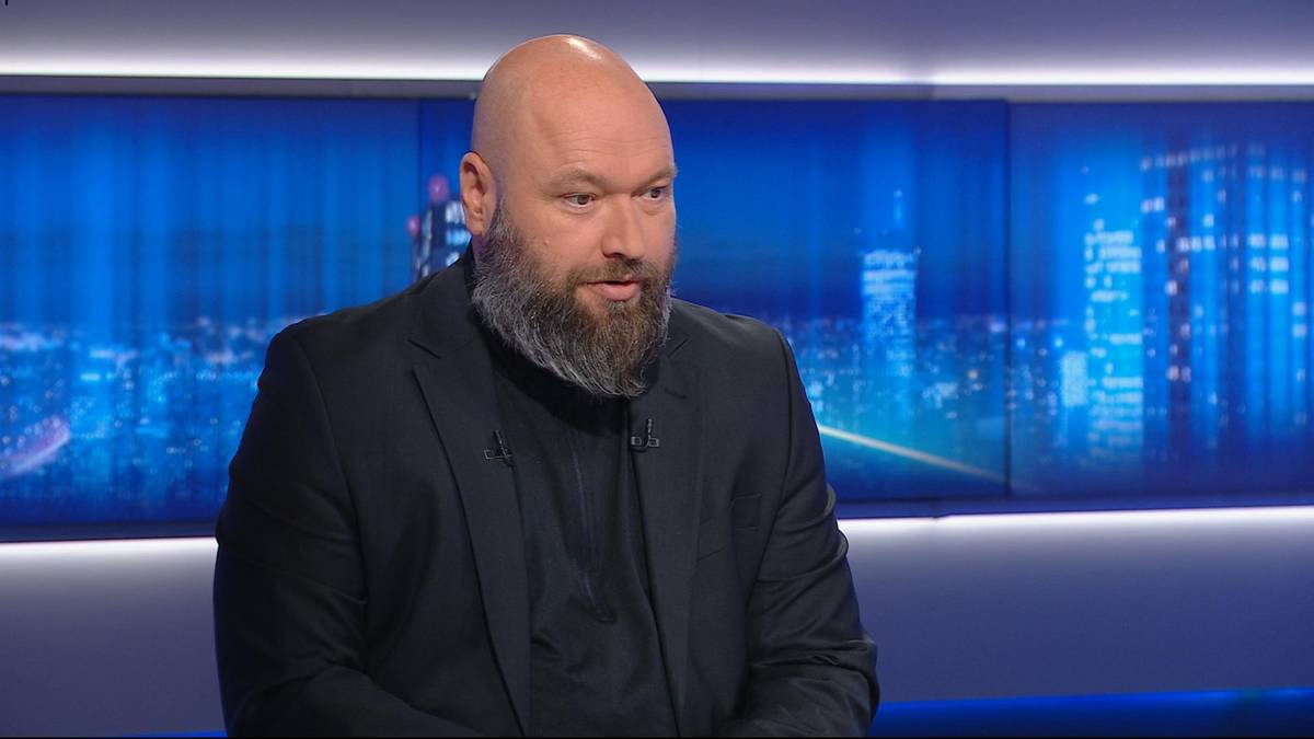 Łysiejący mężczyzna z brodą w ciemnym garniturzie siedzi w studio telewizyjnym na tle niebieskich ekranów.