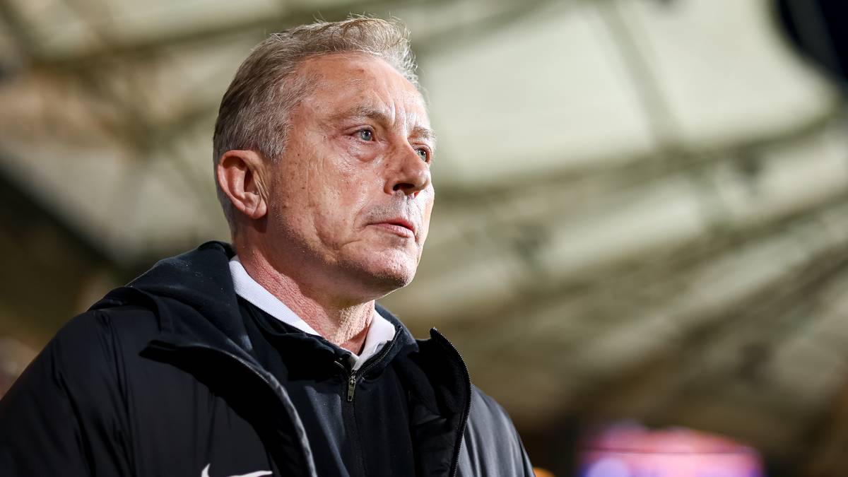 Portret mężczyzny, trenera piłkarskiego, w średnim wieku, patrzącego w bok z zamyślonym wyrazem twarzy. Jest ubrany w sportową kurtkę.