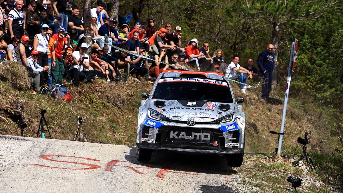Rajdowa Toyota GR Yaris Rally2 z napisem KAJTO na przedniej szybie, pokonująca zakręt na drodze, z kibicami obserwującymi wyścig na zboczu wzg&oacute;rza.