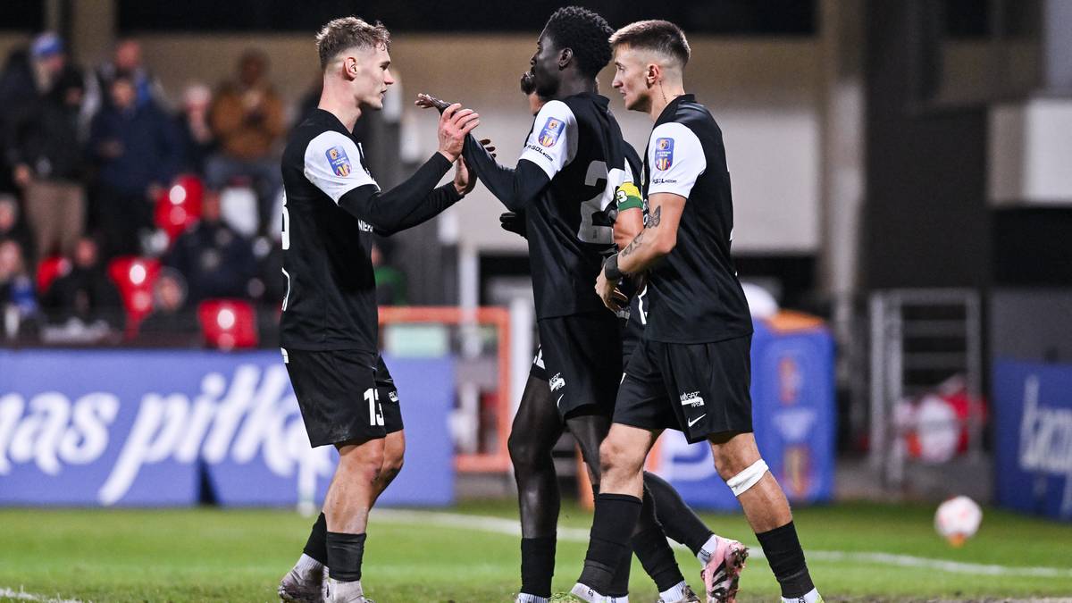 Trzech piłkarzy w czarnych strojach świętuje bramkę na boisku piłkarskim.