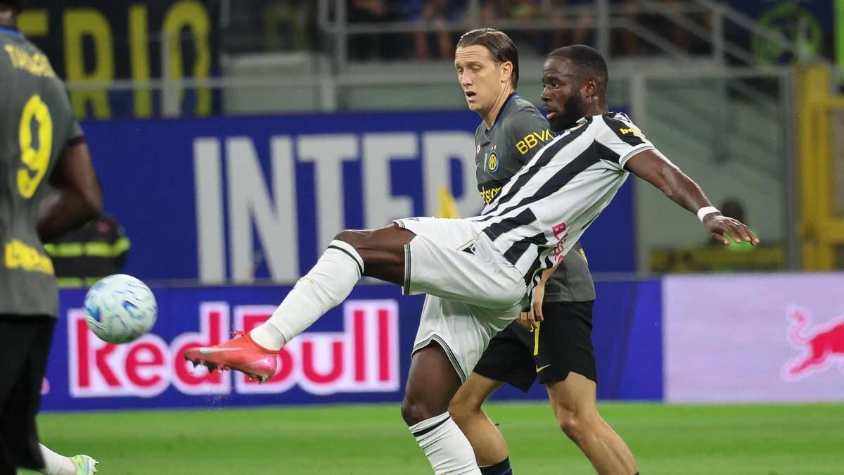 Dw&oacute;ch piłkarzy podczas meczu, jeden w białej koszulce z czarnymi pasami pr&oacute;buje kopnąć piłkę, drugi w ciemnej koszulce pr&oacute;buje go zatrzymać.