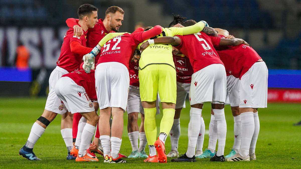 Drużyna piłkarska w czerwonych strojach i jeden zawodnik w żółtym stroju, tworzą zjednoczony krąg na boisku, emanując determinacją i współpracą.