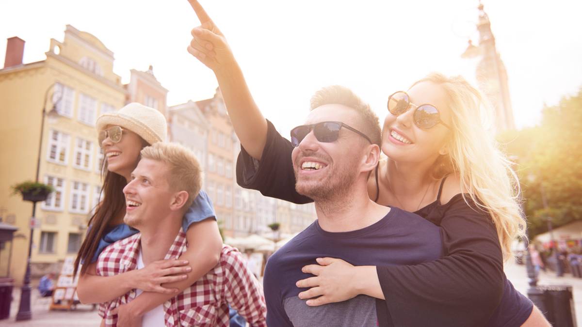 Grupa uśmiechniętych os&oacute;b zwiedzających miasto, jedna z os&oacute;b wskazuje palcem w g&oacute;rę.