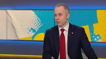 Mężczyzna w ciemnym garniturze, białej koszuli i czerwonym krawacie, siedzący w studiu telewizyjnym.