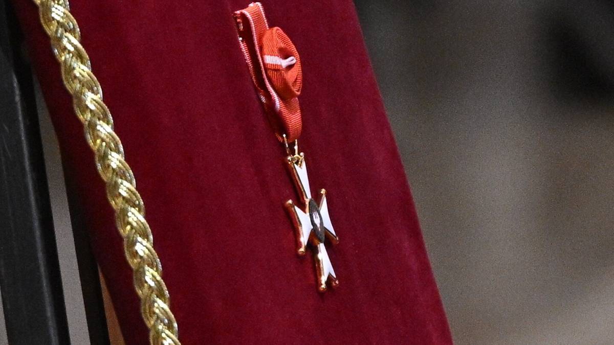 Zbliżenie na ozdobę w kształcie krzyża z białymi ramionami i złotym środkiem, przypiętą do czerwonego materiału z ozdobną złotą plecionką.