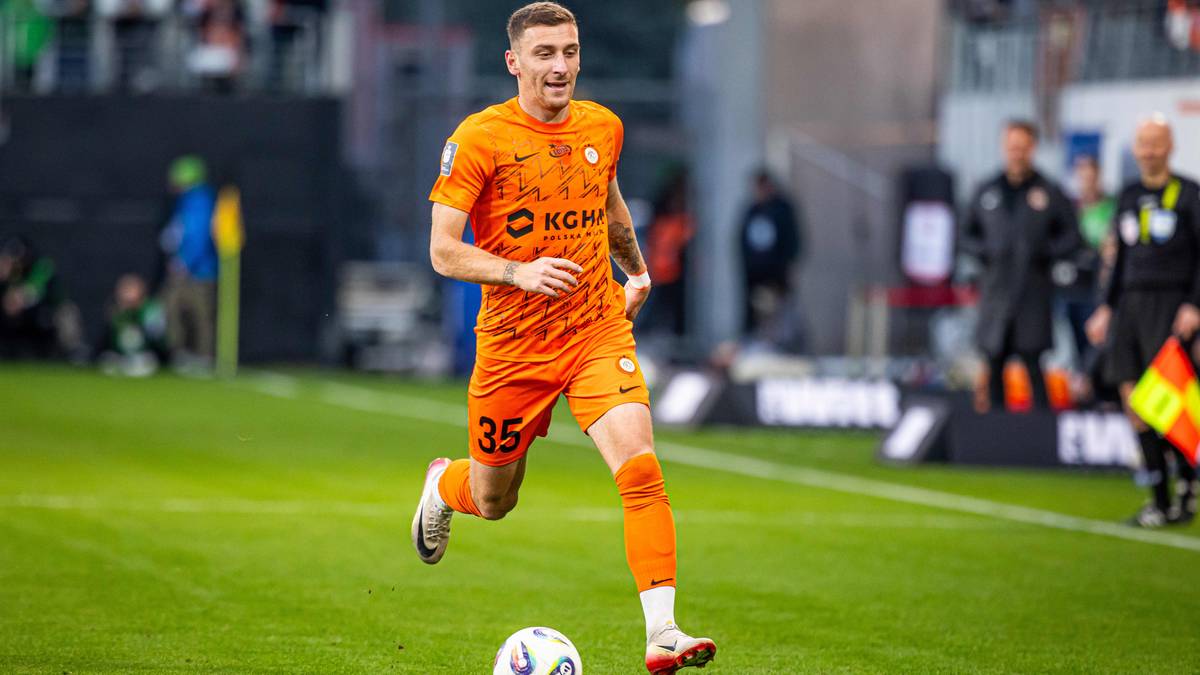 Piłkarz w pomarańczowej koszulce z numerem 35 i białymi pasami na rękawach, biegnie z piłką na zielonym boisku piłkarskim. W tle widać innych zawodnik&oacute;w i kibic&oacute;w.