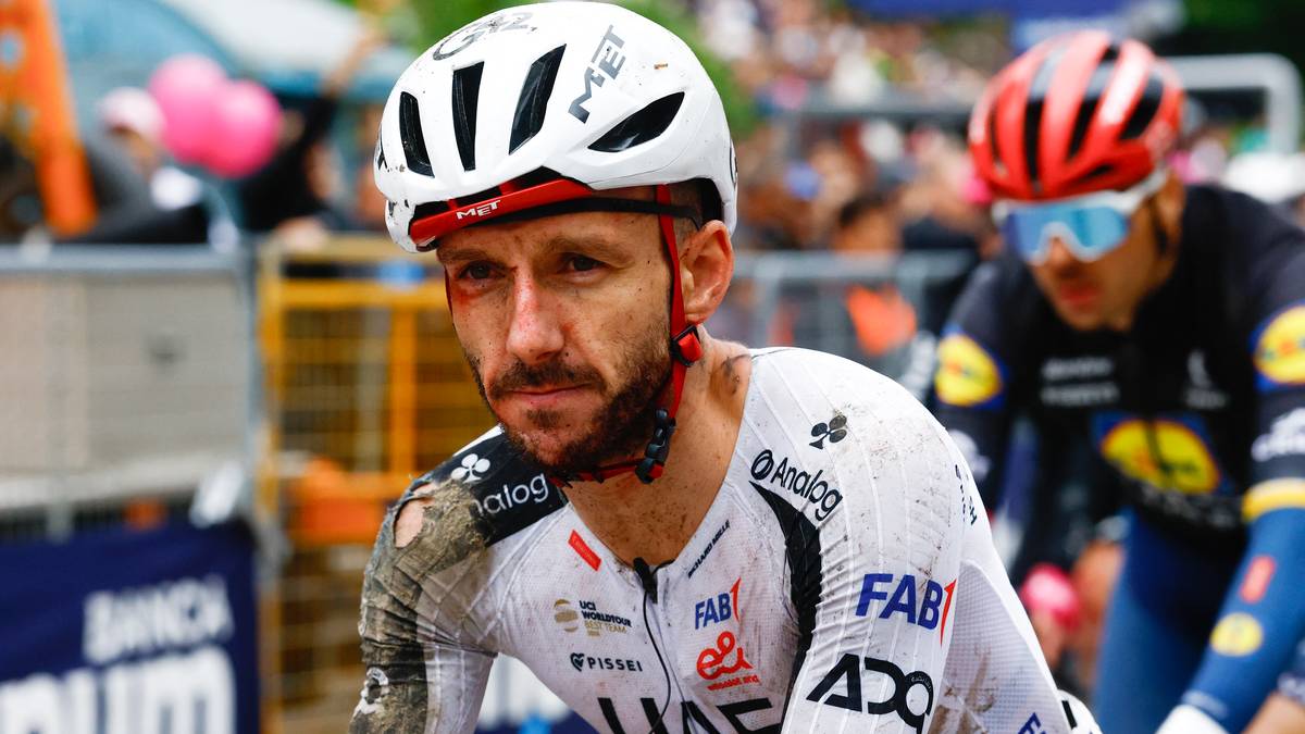 Kolarz w białym stroju z logo drużyny UAE Team Emirates na kasku i koszulce, z zarostem i widocznymi otarciami na twarzy.