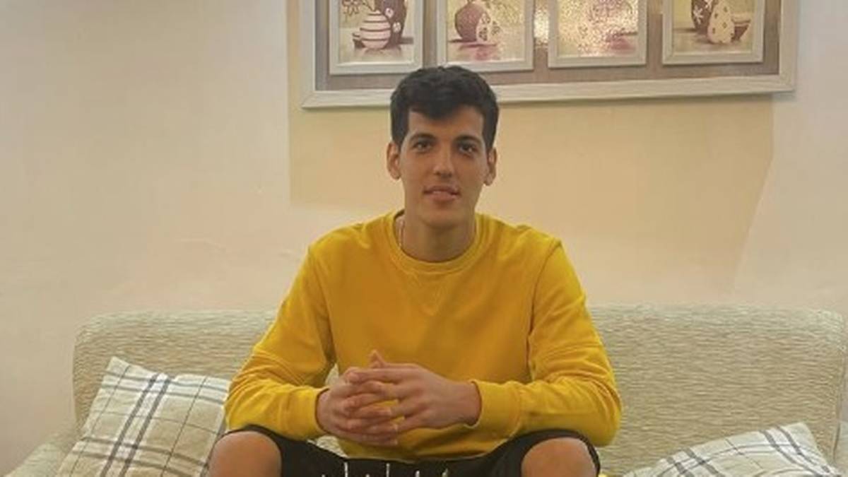 Młody mężczyzna w ż&oacute;łtej bluzie z długim rękawem siedzi na kanapie, ze splecionymi dłońmi na kolanach.