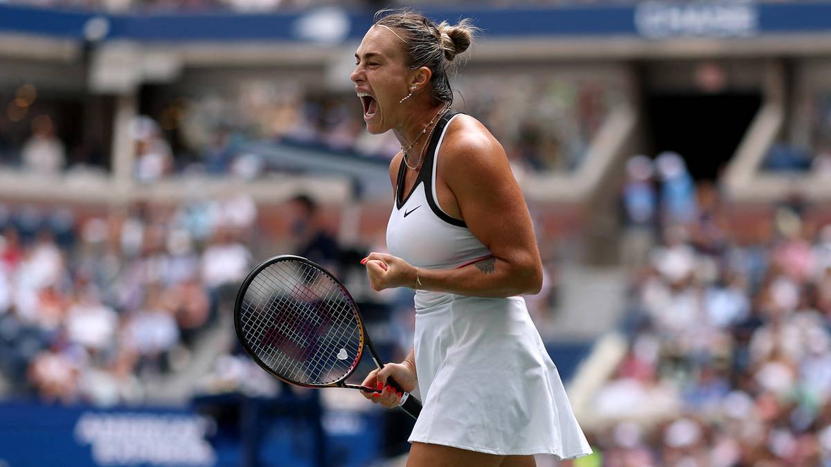 Aryna Sabalenka w białej sukience tenisowej, podnosząc prawą rękę w geście radości, z rakietą tenisową w lewej ręce.