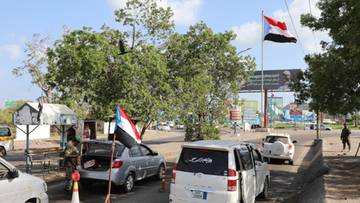 Kilkanaście samochod&oacute;w osobowych i terenowych zaparkowanych przy drodze, niekt&oacute;re z nich z przyczepionymi flagami Jemenu. W tle budynek, drzewa i maszt z flagą Jemenu.