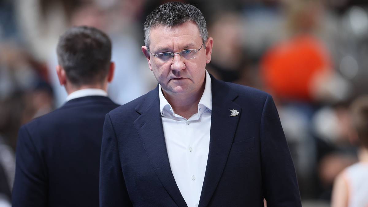 Mężczyzna w garniturze i okularach, z poważną miną, na pierwszym planie.