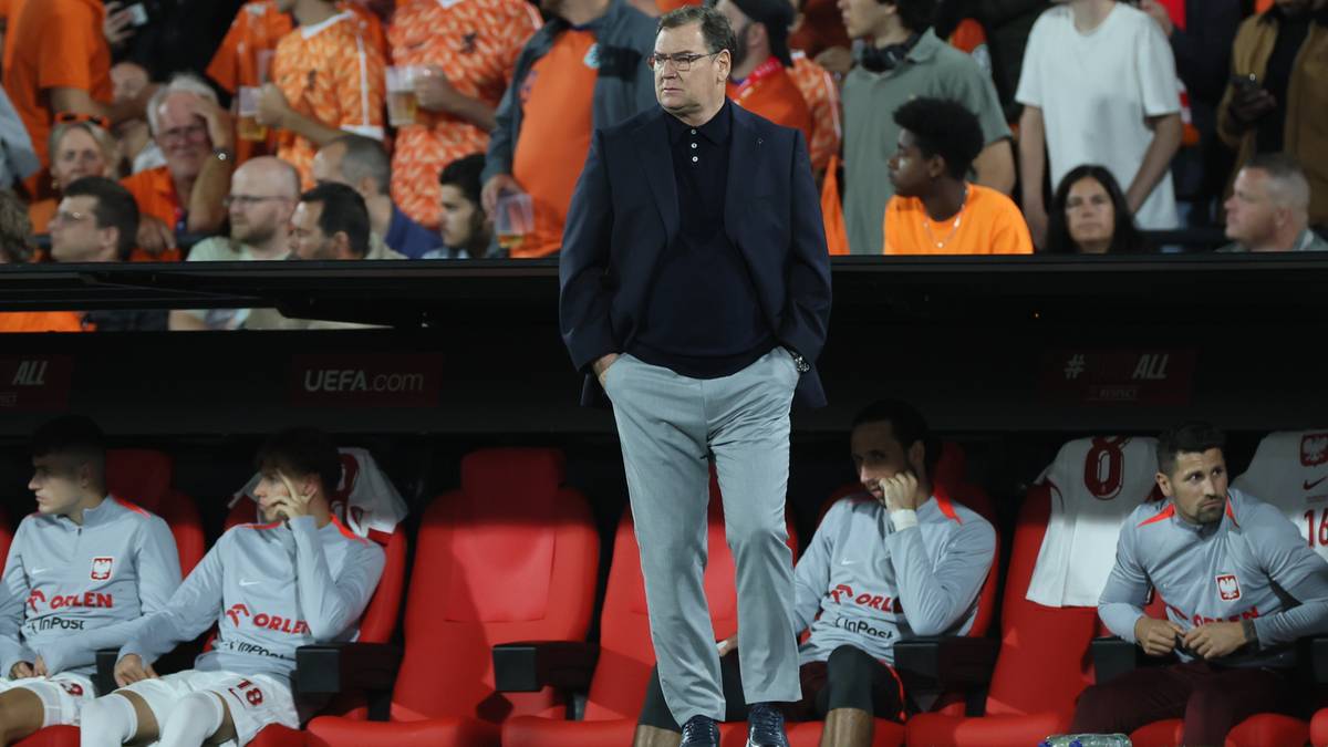 Trener w garniturze stoi na ławce rezerwowych, obserwując mecz. Za nim siedzą piłkarze w szarych bluzach.