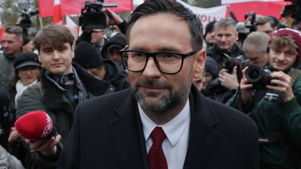 Daniel Obajtek, europoseł PiS, w okularach i garniturze, stoi przed tłumem ludzi i flagami, podczas gdy dziennikarze kierują w jego stronę mikrofony.