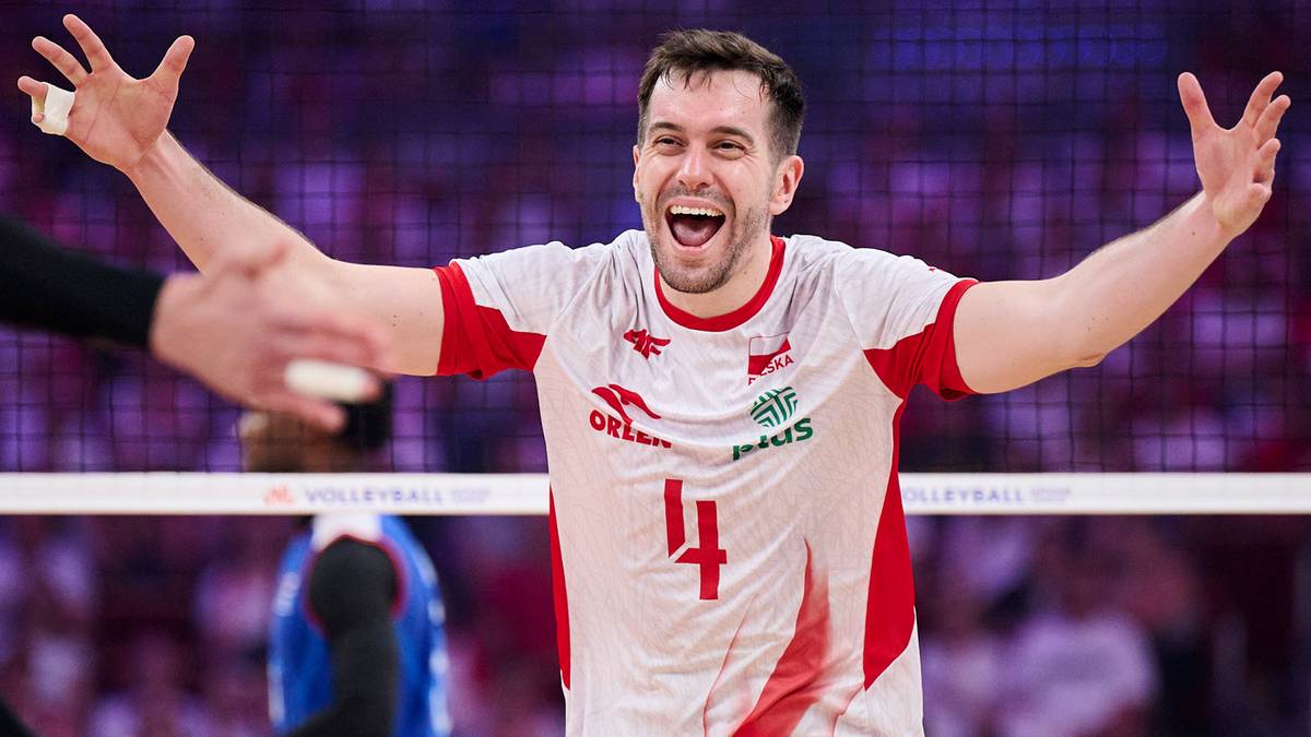 Mężczyzna w białej koszulce z czerwonymi elementami i numerem 4, trzyma otwarte ręce w geście radości, uśmiecha się szeroko. Tło jest rozmyte, widoczna jest siatka do siatkówki.