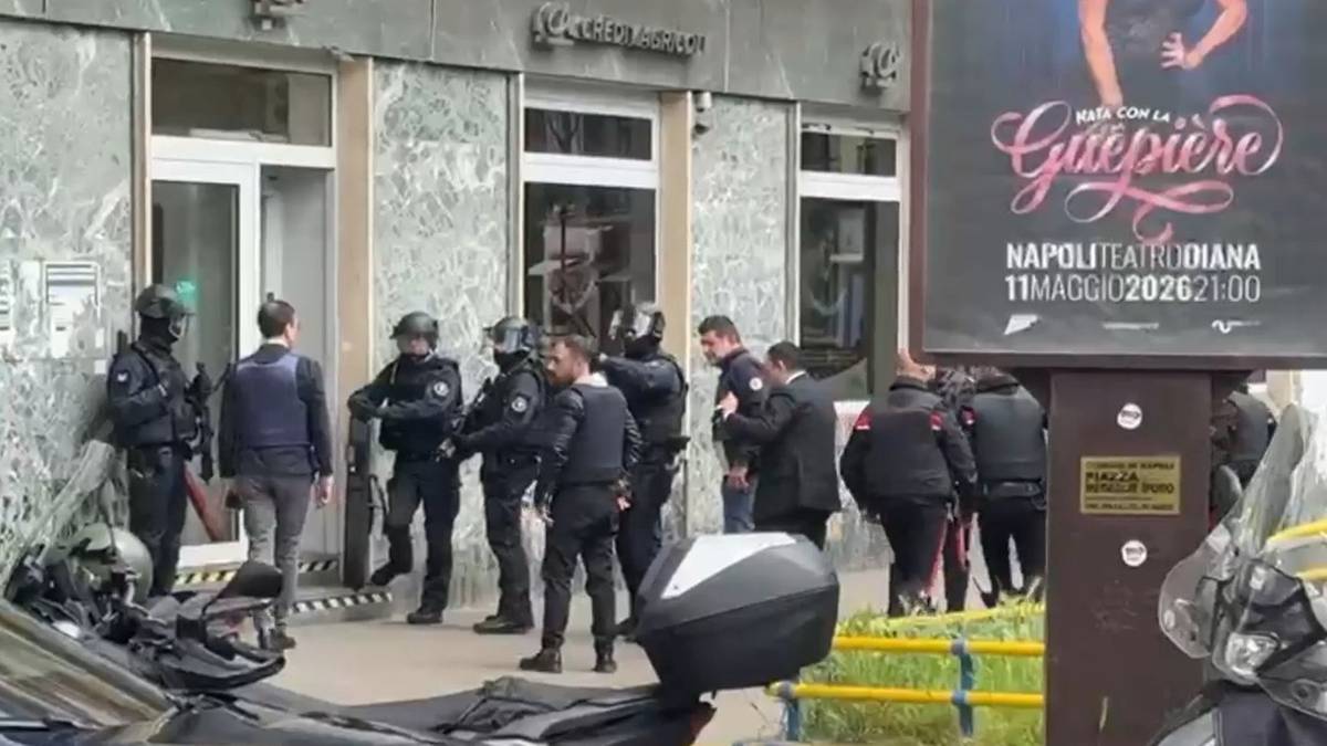Policjanci w pełnym rynsztunku przed zdewastowanym budynkiem banku, z widocznym fragmentem samochodu na pierwszym planie.