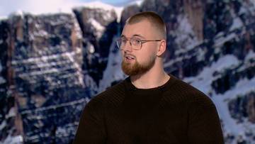 Młody mężczyzna w okularach i z zarostem, ubrany w ciemny sweter, na tle ośnieżonych g&oacute;r.