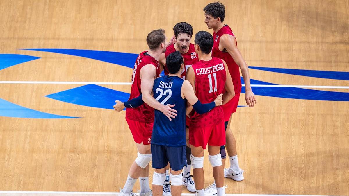 Siatkarze w czerwonych i niebieskich strojach sportowych stoją w kręgu na boisku siatkarskim, dyskutując między sobą.