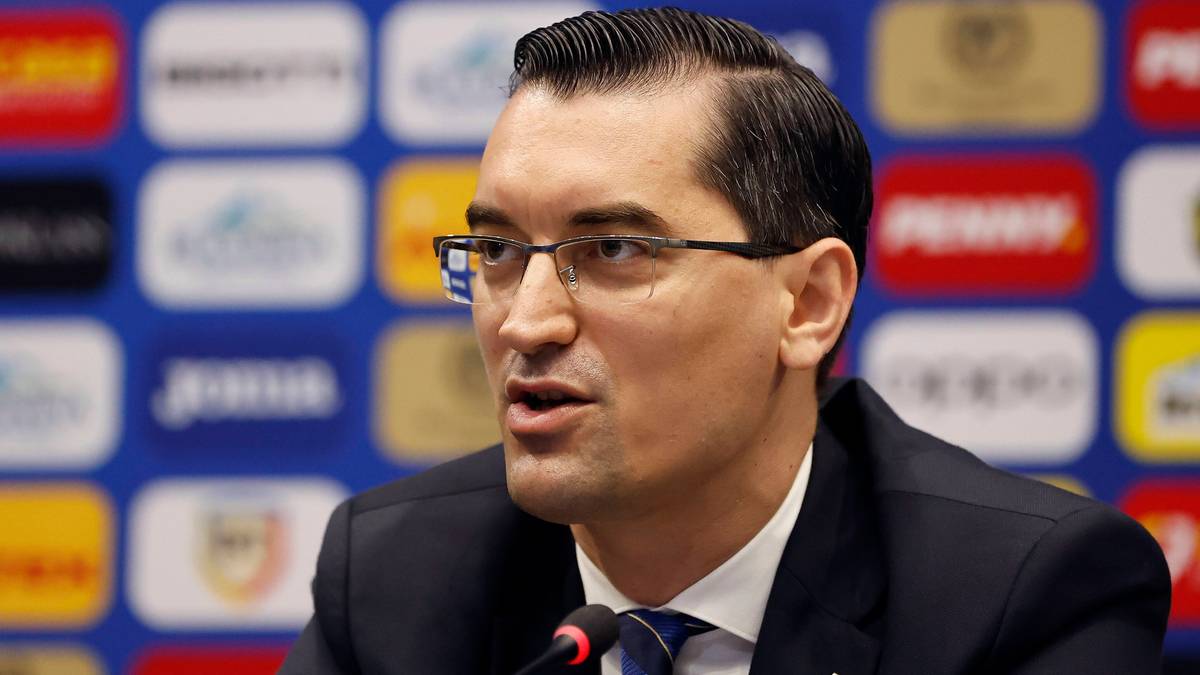 Mężczyzna w okularach i garniturze m&oacute;wi do mikrofonu na tle wielu kolorowych logo.