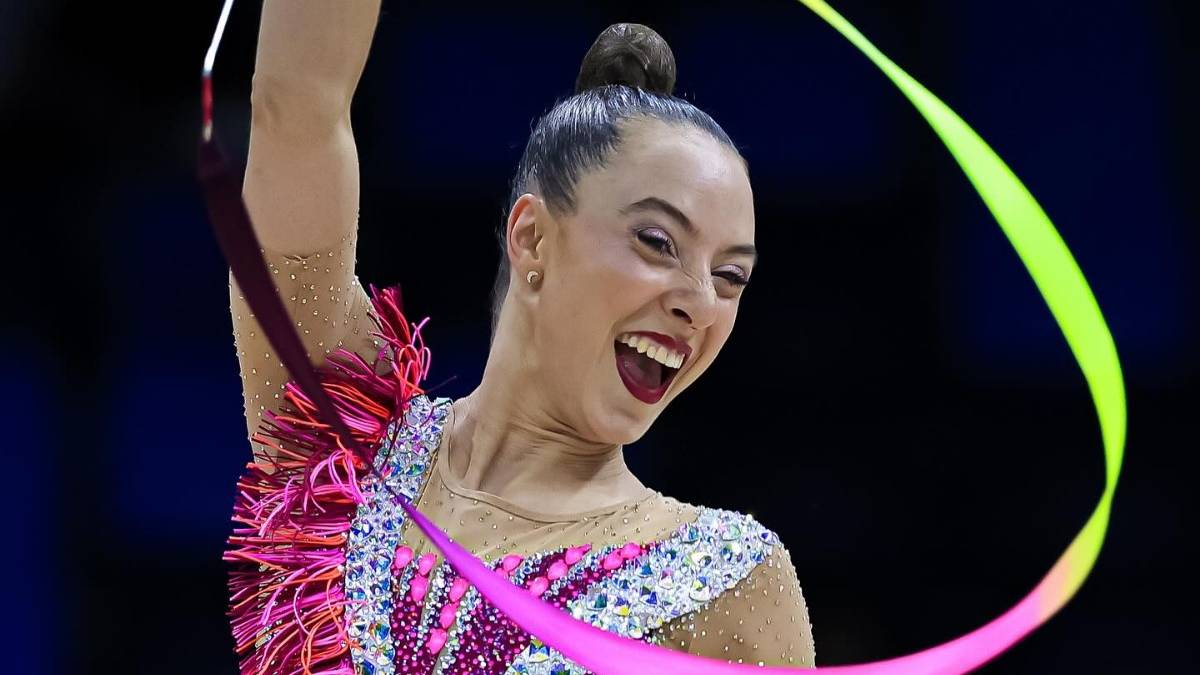 Gimnastyczka artystyczna wykonująca układ z wstążką, z szerokim uśmiechem na twarzy.