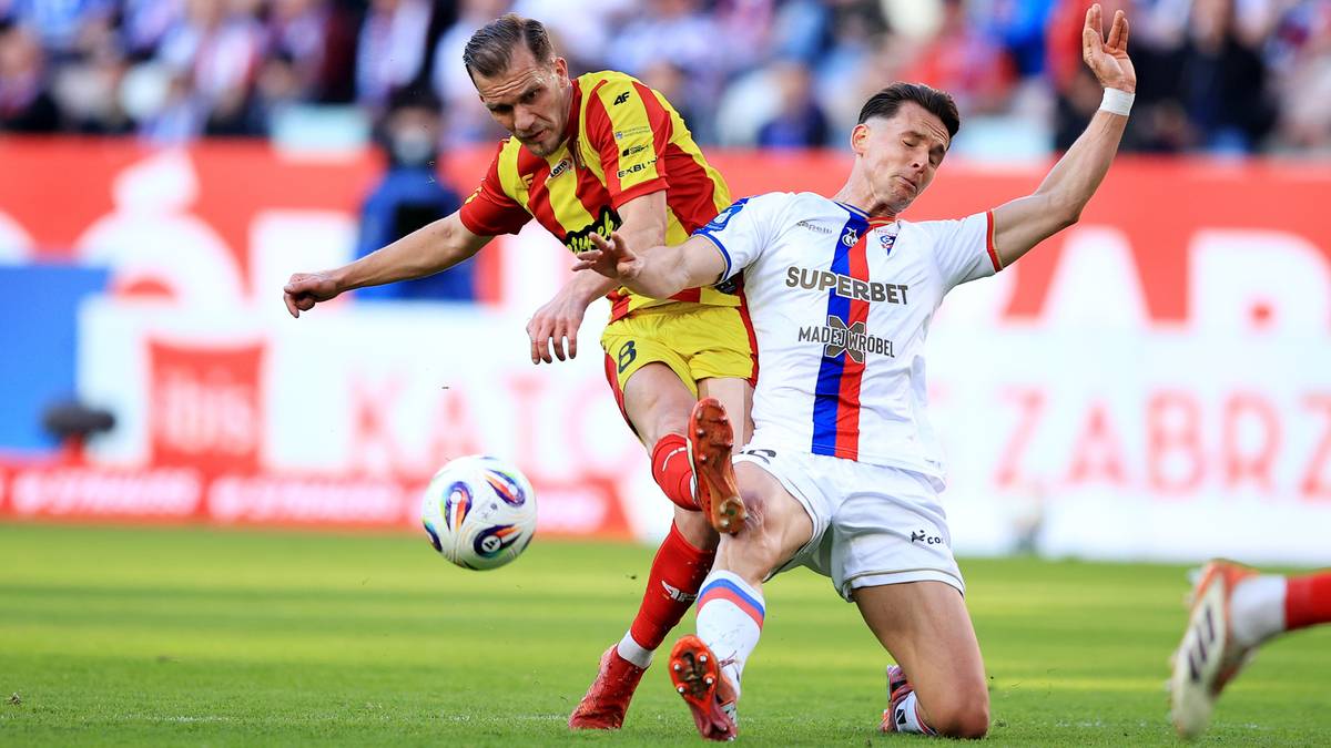 Piłkarz w ż&oacute;łto-czerwonym stroju pr&oacute;buje odebrać piłkę zawodnikowi w białym stroju z czerwono-niebieskimi pasami.