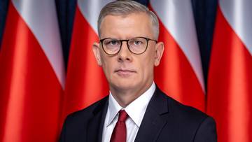 Portret Sławomira Cenckiewicza, szefa Biura Bezpieczeństwa Narodowego, z okularami, w garniturze i krawacie, na tle białych i czerwonych pas&oacute;w polskich flag.