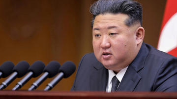 Portret Kim Dzong Una przemawiającego przy podium z mikrofonami.