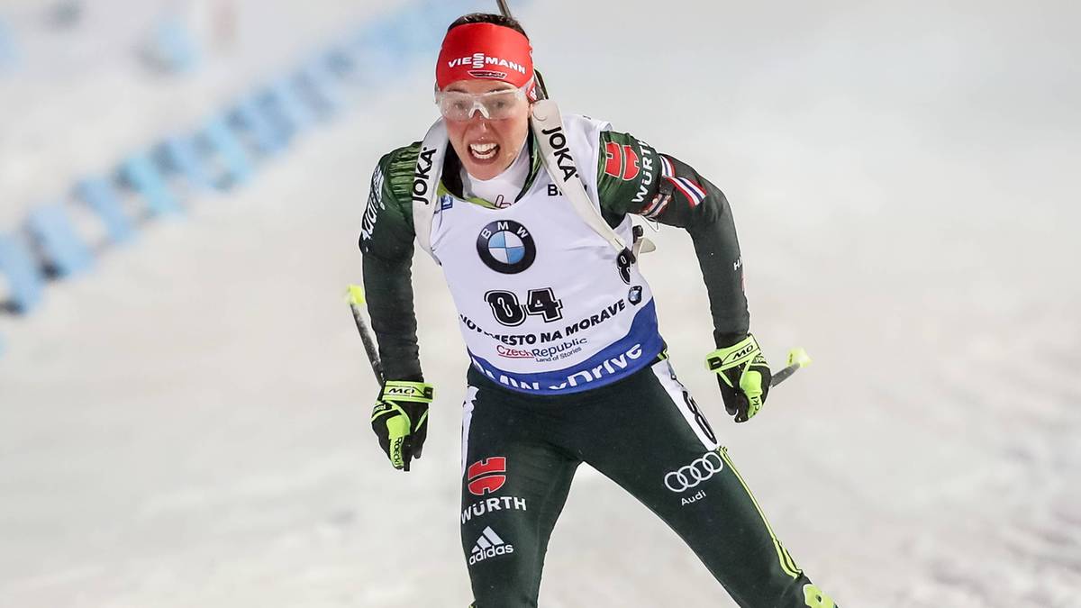 Biathlonistka w kombinezonie sportowym i goglach, z karabinem na plecach, zmierza po śniegu.