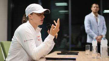 Iga Świątek m&oacute;wi do mikrofonu, wskazując palcami, w białej czapce i bluzie sportowej.