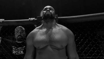 Czarno-białe zdjęcie zawodnika MMA Geronimo Dos Santosa stojącego w klatce, patrzącego przed siebie z podniesioną głową. W tle widoczna jest sylwetka innej osoby.