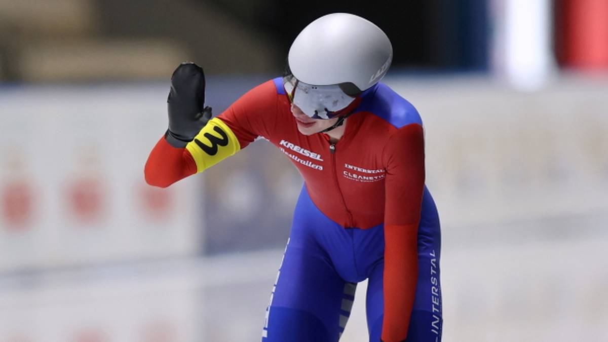 Zawodniczka w czerwono-niebieskim kombinezonie panczenowym, z białym kaskiem i goglami, unosi rękę w geście pozdrowienia. Na lewym ramieniu ma ż&oacute;łtą opaskę z numerem 3.