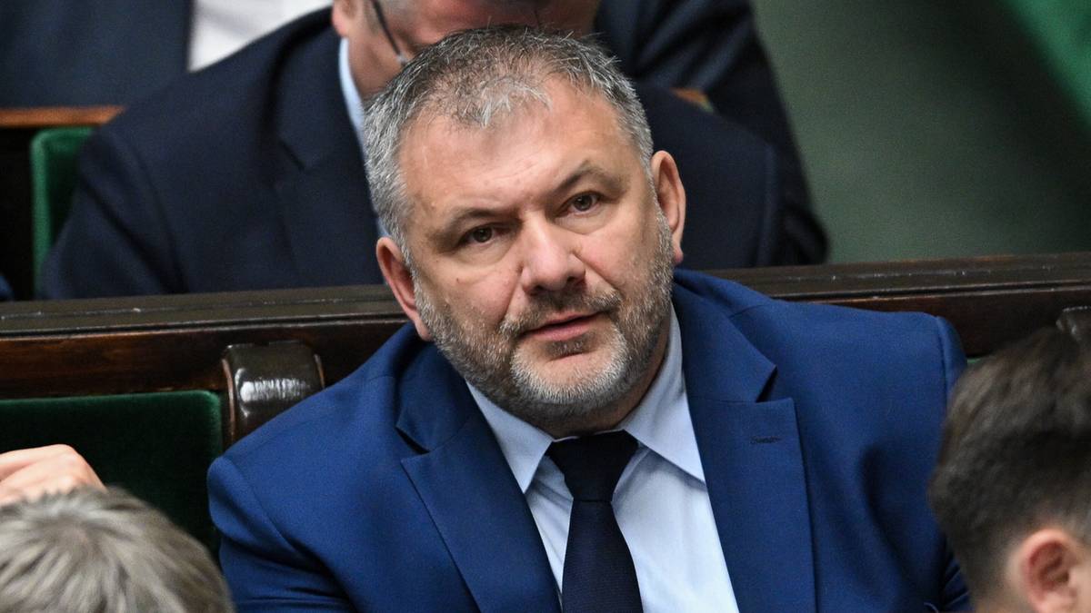 Mężczyzna w niebieskim garniturze i krawacie siedzi w pierwszym rzędzie.