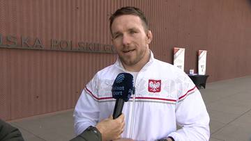 Tadeusz Michalik, polski zapaśnik, w białej bluzie z biało-czerwonymi detalami, udziela wywiadu z mikrofonem przed budynkiem z napisem "POLSKIE GO".