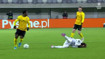 Piłkarz w żółtej koszulce biegnie z piłką po boisku, podczas gdy drugi piłkarz w białym stroju leży na ziemi.