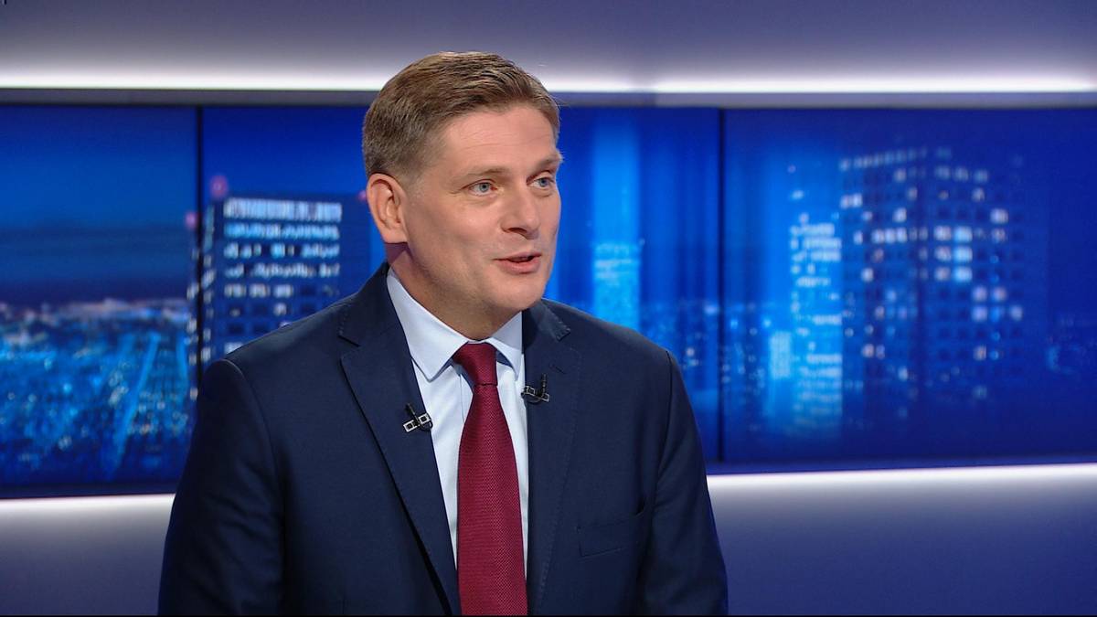 Mężczyzna w garniturze i krawacie m&oacute;wi przed ekranem z nocnym pejzażem miasta