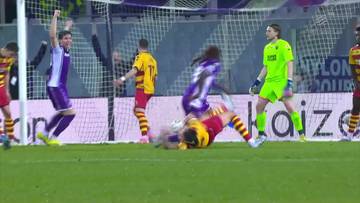 Piłkarz w czerwono-żółtym stroju leży na murawie po zderzeniu z przeciwnikiem. Obok niego stoją inni zawodnicy w fioletowych i żółtych koszulkach oraz bramkarz w zielonym stroju.