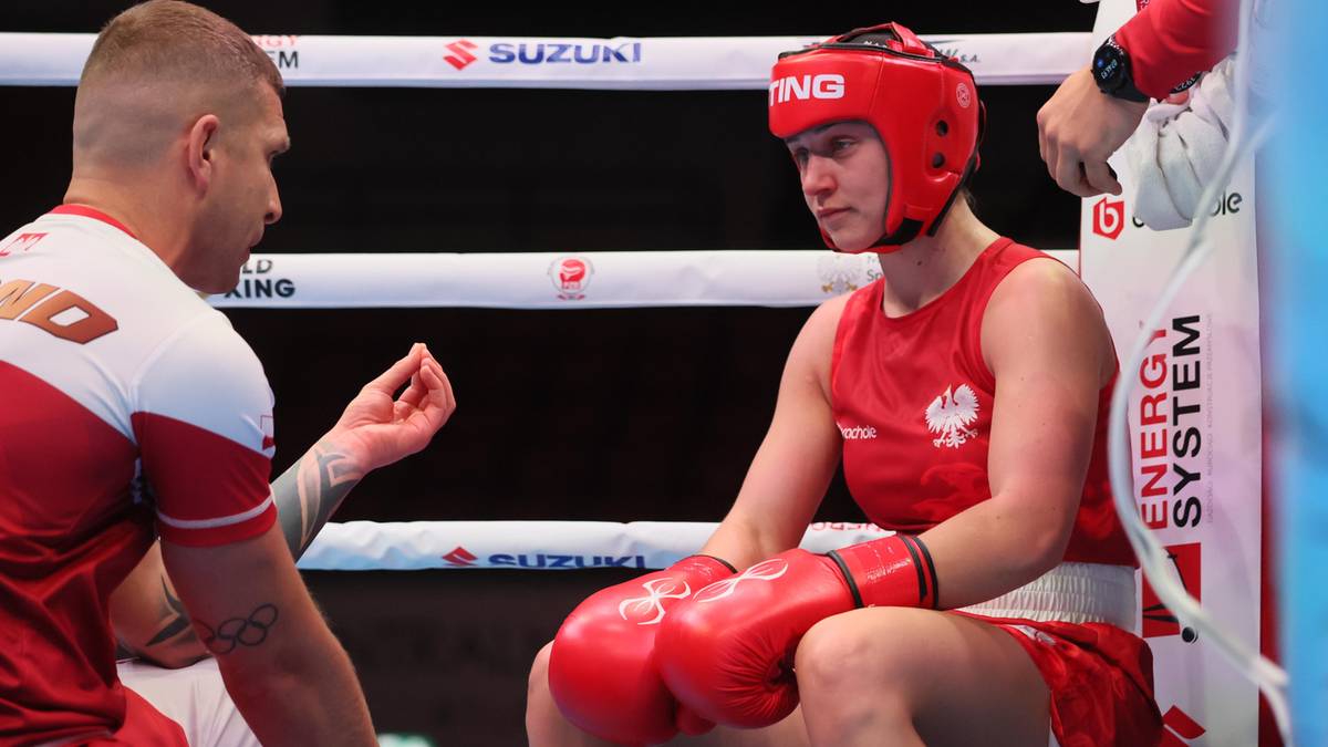 Trener bokserski w białej koszulce z czerwonymi elementami zwraca się do siedzącej bokserki w czerwonym stroju i kasku bokserskim.